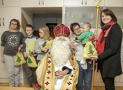 In the center of the picture is St. Nicholas. To his right are two girls and a boy. All three have a St. Nicholas bag in their hands. To the left of St. Nicholas stands a woman who has a small child on her arm. Next to her is Stefanie Rud, who is also holding a St. Nicholas bag and the holy staff in her hands.