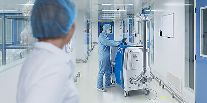 A large bright hallway. On the left is the torso of a person dressed in white with protective hood. In the background, a man in a blue protective suit, protective hood, blue gloves and white mouth guard is standing at a white-blue box on rollers. The box goes up to the person's chest. On the front of the box you can see ISU. Above the box is a tablet on which the person is working. On the side of the box, at the bottom, there is a tube that protrudes up along the box. The tube is about one and a half times as long as the box is high. At the end of the tube is a plate where six smaller curved tubes protrude. 