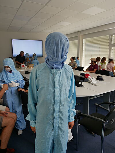 In a large room, a table at which children are sitting can be seen in the background. In the foreground is a child wearing a cleanroom suit with his cap pulled over his face. Behind him is also a child in a cleanroom suit with a mask and gloves.