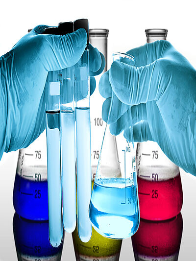 In the background are Erlenmeyer flasks with blue, yellow and red liquid. In the foreground are two hands with blue protective gloves. On the left are three test tubes with blue liquid, on the right is an Erlenmeyer flask with blue liquid.