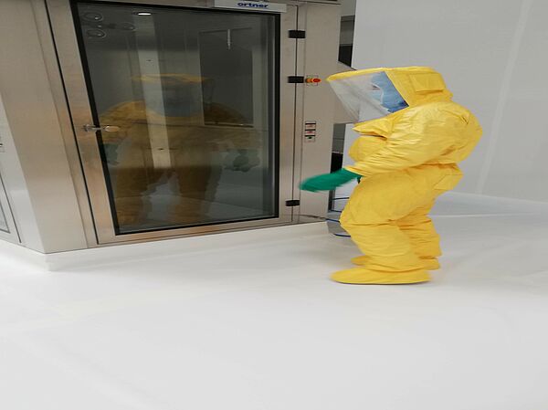 A person wearing a yellow protective suit is standing in front of a personal wet shower. She is also wearing a hard hat, which is transparent at the front.