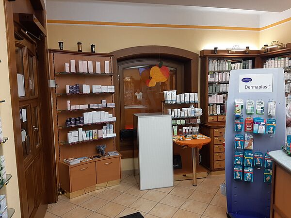  In a pharmacy room, on the left, there is a low box on which brochures are lying. Above it, five shelves are mounted on the wall on which different small boxes and cream tubes are placed. On the right side in the background is a corner shelf. The lower part consists of five drawers. Above are again five shelves filled with small boxes and cream tubes. In the foreground is a display on which four packs of different plasters hang in five rows. Between the two shelves in the background is a table on the right, which has a small shelf at the back end. On four levels are small boxes and tubes of cream. To the left is a small box that has a gray frame. The front consists of a fine white grid.