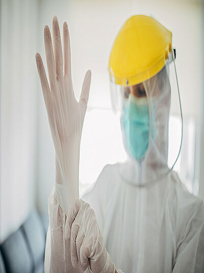 A person wearing white protective clothing, mouth guard and helmet with transparent protective shield is putting on a protective glove.
